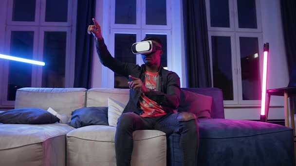 Good-looking serious young dark-skinned man in virtual reality headset sitting on the sofa in the room and moving hands on imaginary screen - Video