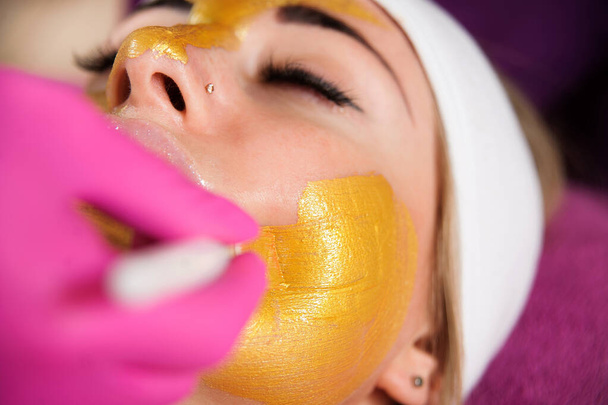 Cosmetisch masker. De schoonheidsspecialiste brengt een gouden masker aan op het gezicht van een vrouw. Mooie vrouw in een schoonheidssalon tijdens gezichtsbehandeling - Foto, afbeelding