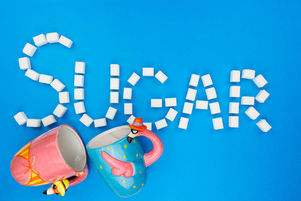 Sugar cubes and two cups on an isolated blue background making up a word Sugar. Pink and blue cups with white cubes of sugar isolated on blue background. Concept word sugar made up of cubes of sugar. - Photo, Image