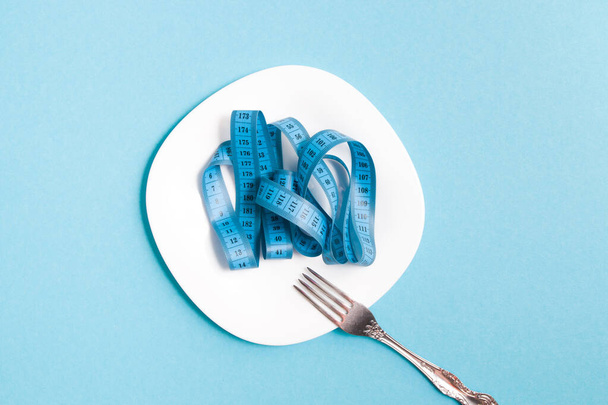 blue measuring tape on a white plate and fork, blue background, opium seats, top view, diet concept, weight loss and fasting - Φωτογραφία, εικόνα