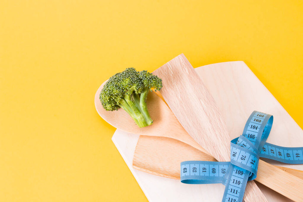 green raw broccoli on wooden kitchen appliances tied up with a blue measuring tape bow, yellow background, copy space, diet concept, eco friendly life style - Fotoğraf, Görsel