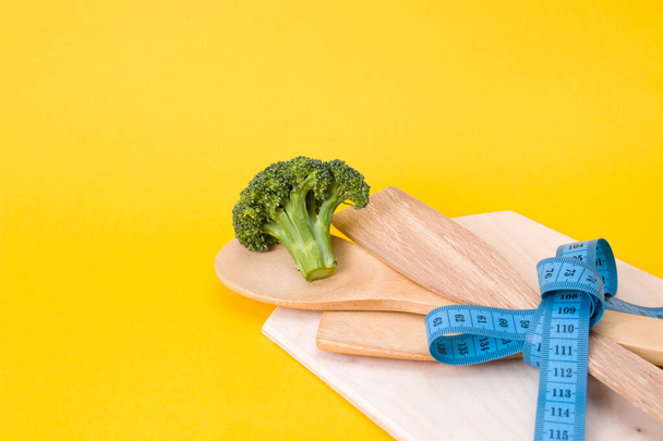 green raw broccoli on wooden kitchen appliances tied up with a blue measuring tape bow, yellow background, copy space, diet concept, eco friendly life style - Fotó, kép