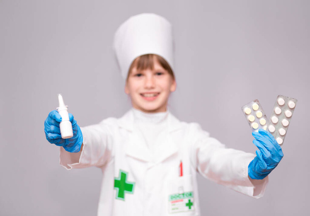 Улыбающаяся маленькая девочка в медицинской форме с носовым спреем и таблетками для здоровья. Врач рекомендует спрей для носа
 - Фото, изображение