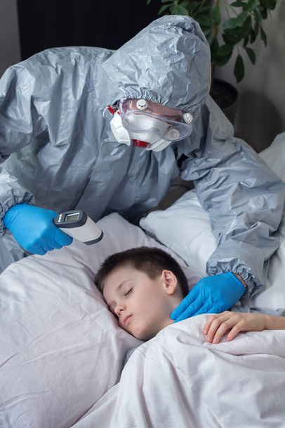 Egy beteg iskolás fiú fekszik az ágyban. Egy védőruhát és gumikesztyűt viselő személy lézerhőmérővel méri a gyermek hőmérsékletét. A másik kezével egy beteg ember pulzusát méri. Segíts az embereknek a karanténban.. - Fotó, kép