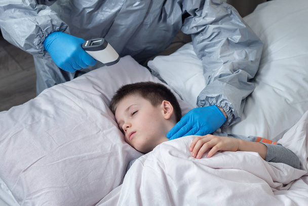 A sick school-age boy is lying in bed. A person wearing a protective suit and rubber gloves measures the child's temperature with a laser thermometer. With the other hand, he measures the pulse of a sick person. Help people in quarantine. - Foto, afbeelding