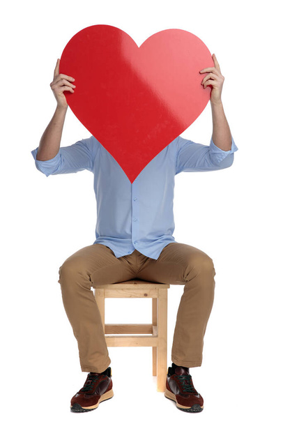 Young casual man covering his face with heart shape while sitting on a chair on white studio background - Foto, Imagem