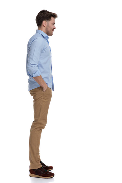 Side view of confident casual man holding both hands in pockets while wearing shirt and standing on white studio background - Photo, Image