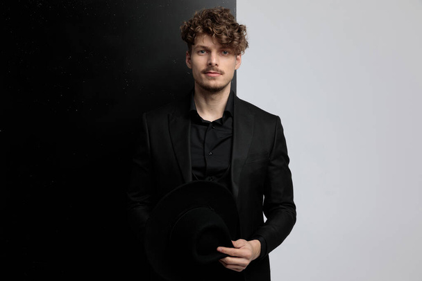 Takım elbise giyerken elinde şapka tutan ve siyah beyaz stüdyo arka planında duran genç bir manken. - Fotoğraf, Görsel