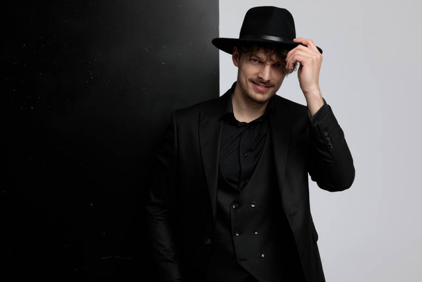 Modelo de moda positiva fixando seu chapéu e saudação enquanto vestindo terno e de pé no fundo do estúdio preto e branco
 - Foto, Imagem