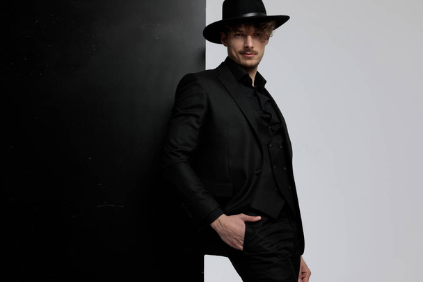 Charmant mode model poseren met zijn hand in zijn zak tijdens het dragen van pak en hoed, staande op zwart-wit studio achtergrond - Foto, afbeelding