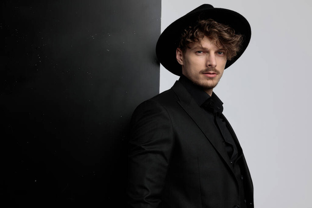 Мотивированная модель с уверенностью смотрит вперед, надевая костюм и шляпу, стоя на черно-белом фоне студии
 - Фото, изображение