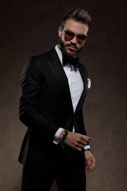 Tough fashion groom looking forward while wearing suit and sunglasses, standing on a wallpaper studio background - Fotografie, Obrázek