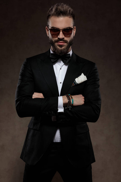 Tough fashion groom holding his hands crossed while wearing suit and sunglasses, standing on a wallpaper studio background - Foto, afbeelding