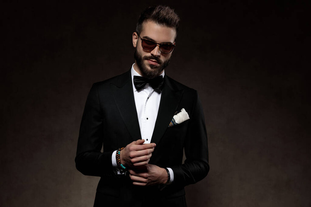 Positive fashion groom adjusting his sleeve and smiling while wearing sunglasses and suit, standing on a wallpaper studio background - Fotografie, Obrázek