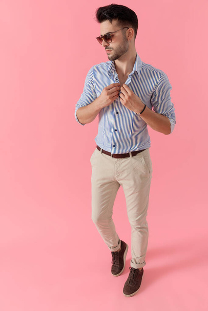 Pensive smart casual man unbuttoning his shirt wearing sunglasses, walking on pink studio background - Foto, afbeelding