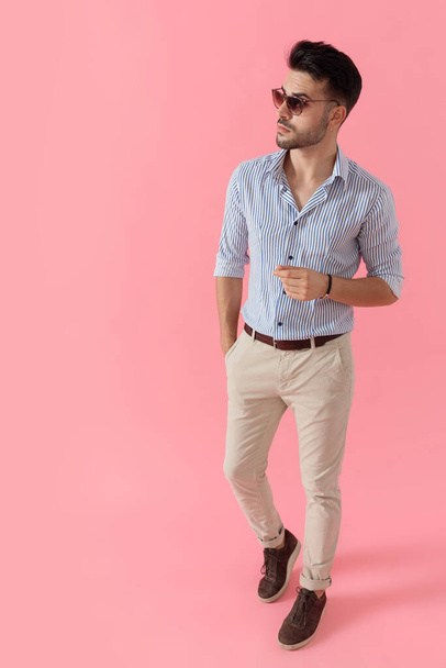 gretige slimme casual man weg te kijken met de hand in zak terwijl het dragen van shirt en zonnebril, lopen op roze studio achtergrond - Foto, afbeelding