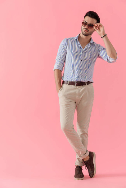 Attractive smart casual man looking away curiously and fixing his sunglasses while wearing shirt, standing on pink studio background - Photo, image