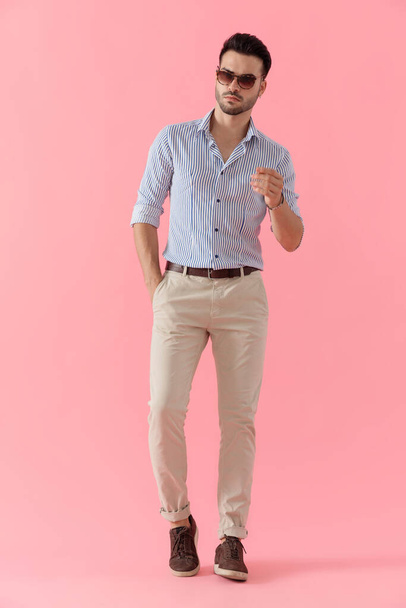 Cool smart casual man holding hand in pocket while wearing shirt and sunglasses, walking on pink studio background - Фото, изображение