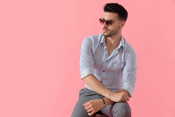 Homme décontracté intelligent pensif regardant loin tout en portant chemise et lunettes de soleil, assis sur un tabouret sur fond de studio rose
 - Photo, image