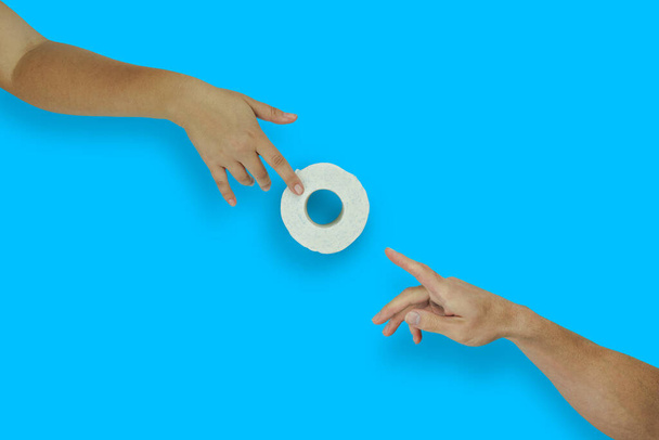 Женская рука дает рулон туалетной бумаги для мужской руки на синем фоне, вид сверху, плоский узор. Покупка паники из-за вспышки коронавируса. Шаловливая метафора Адама Микеланджело
 - Фото, изображение