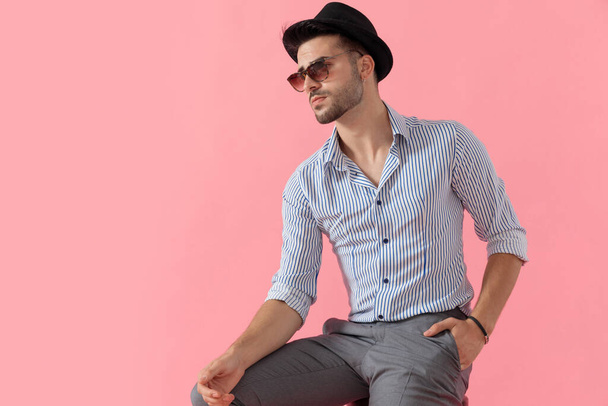 Hoopvolle slimme casual man die wegkijkt terwijl hij hoed, zonnebril en shirt draagt, zittend op een kruk op roze studioachtergrond - Foto, afbeelding