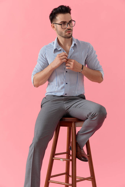 Gözlüklü, pembe stüdyo arka planında bir taburede otururken gömleğinin düğmelerini açan hevesli, sıradan bir adam. - Fotoğraf, Görsel
