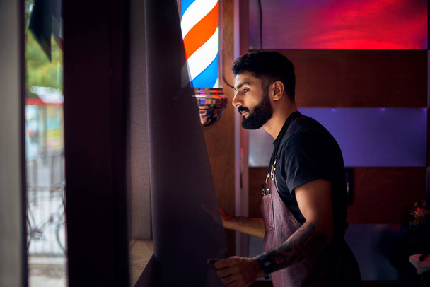 Young hairdresser at his hairdresser. a man with a beard is standing by the window and looking at the street. - 写真・画像