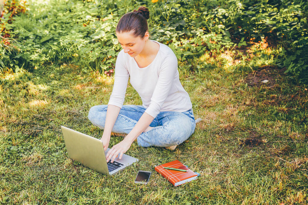 Mobile Office. Freelance business concept. Young woman sitting on green grass lawn in city park working on laptop pc computer. Lifestyle authentic candid student girl studying outdoors - Photo, Image