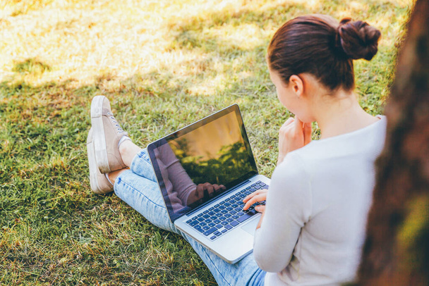 Мобільний офіс. Концепція вільного бізнесу. Молода жінка сидить на зеленому газоні в міському парку, працюючи на комп'ютері для ноутбуків. Стиль життя справжня відверта студентка, яка вивчає на відкритому повітрі
 - Фото, зображення