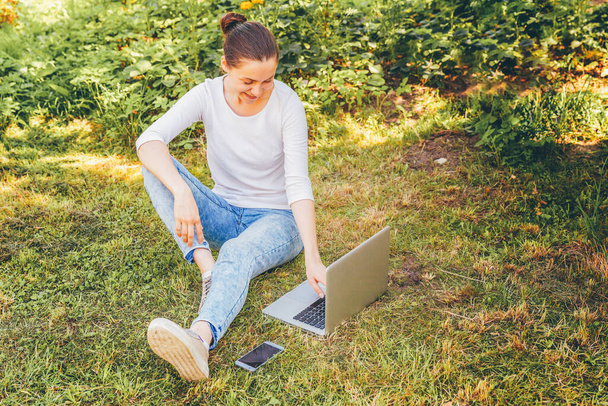 Serbest iş kavramı. Dizüstü pc bilgisayar üzerinde çalışan Şehir Parkı yeşil çim Çimenlikte oturan genç kadın. Yaşam tarzı otantik samimi öğrenci kız açık havada okuyor. Mobil ofis - Fotoğraf, Görsel