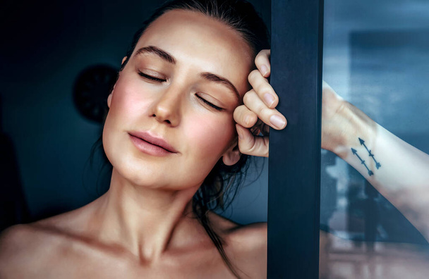 Portrait of a beautiful woman in a dreamy mood with closed eyes standing near the window at home, genuine beauty of a young female - Foto, afbeelding