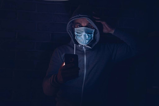 スマートフォンでcovid-19コロナウイルスに関する情報を読む男を怖がらせました。コロナウイルスcovid-19流行中の医療マスクの男. - 写真・画像