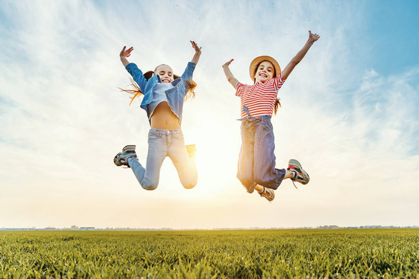 D'en bas de joyeuses petites sœurs en vêtements décontractés s'amuser et sauter avec les bras levés dans un champ vert en été evenin
 - Photo, image
