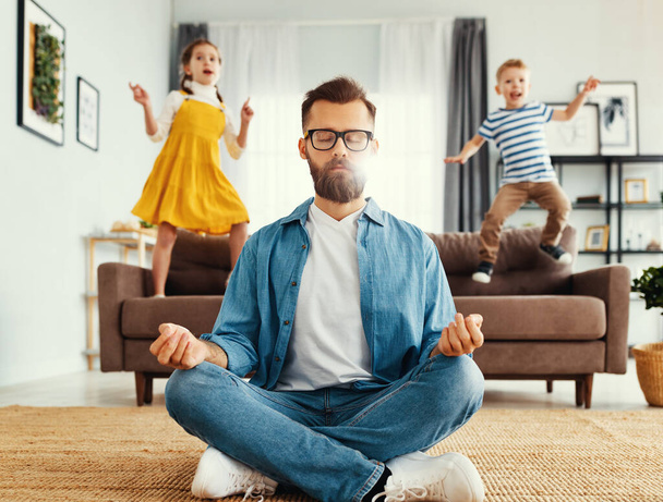 Tranquil young man in casual wear sitting on floor in lotus position and meditating while kids having fun and jumping on sofa at hom - Fotó, kép