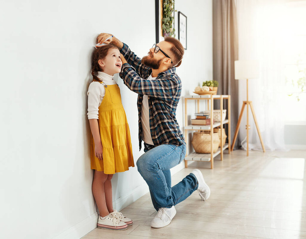 Side view of young man in casual clothes measuring height of little daughter next to white wall in light living roo - Фото, зображення