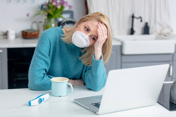 senhora loira em máscara com xícara de bebida, termômetro e computador portátil na cozinha
  - Foto, Imagem