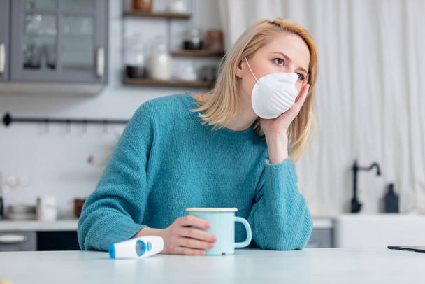 Blonde lady in mask with cup of hot tea and thermometer at kitchen table  - Foto, immagini