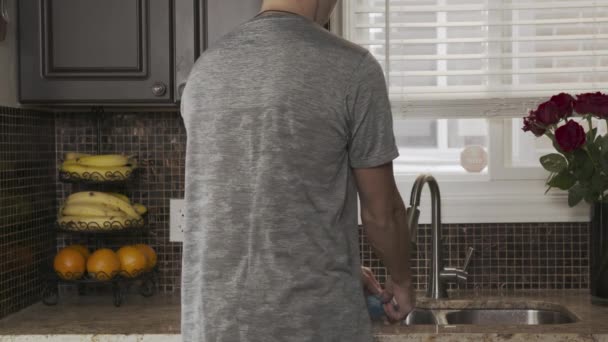 giovane uomo attraente in una t-shirt grigia sta con la schiena alla macchina fotografica e lava i piatti o le mani in una bella cucina pulita alla moda nel lavandino. Un ragazzo lava verdure e frutta nel
 - Filmati, video