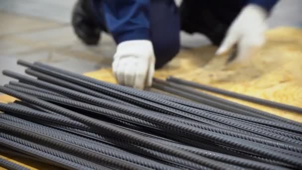 One man worker wear blue overalls, protective white gloves, takes, shifts long bars of rebar lying on floor in neat piles on a wooden pallet standing next to him. Close up view of employee hands. - Záběry, video