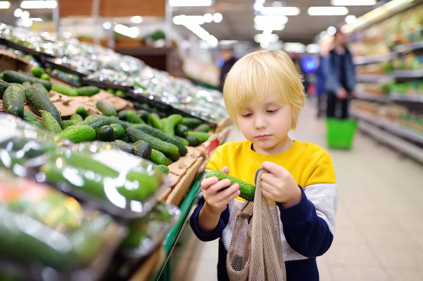 Little boy choosing organic cucumbers and put into mesh shopping bag in food store or supermarket. Healthy lifestyle for family with kids. Concept of zero waste, recycling and protection environment. - Photo, Image