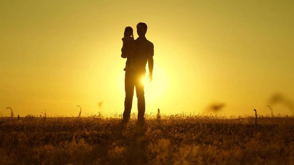 Pai feliz com uma filha pequena nos braços ao pôr-do-sol. pai e bebê caminham no parque. família feliz com a criança estão andando no campo ao pôr do sol. conceito de uma família feliz e infância
. - Foto, Imagem
