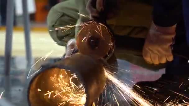 Male employee wears protective blue special uniform, white gloves on hands. Man cuts rusty metal pipe with grinder close up. Sparks fly in all directions. Next worker help, hold, turns pipe for slide. - Imágenes, Vídeo