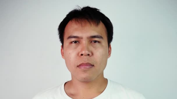 Close-up portret van zieke Aziatische man in wit T-shirt blaast een neus over grijze achtergrond. Mannelijk gezicht met griepsymptomen.  - Video