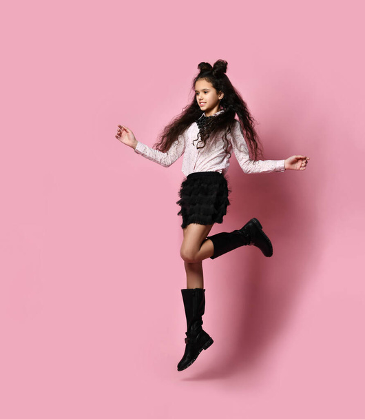 Teenage female with long hair, in white blouse, black skirt and boots. Smiling and jumping against pink background. Full length - 写真・画像