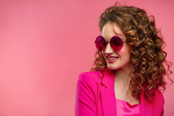 Close up fashion studio portrait of young beautiful women with curly hair.Nice girl with wavy hairstyle,bright make up,stylish eyeglasses isolated over pink background. Beauty and hair care concept - Fotoğraf, Görsel