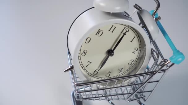 Reloj despertador blanco en un carro de supermercado decorativo sobre un fondo claro. El concepto de descuentos y ahorros financieros
. - Metraje, vídeo