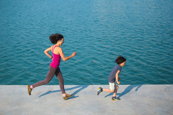 Jogging-Junge mit Mama. Ein Kind läuft mit seiner Mutter auf Destillation. Eine Frau und ihr Sohn laufen die Uferpromenade entlang. Sportfamilie. Gesunder Lebensstil. - Foto, Bild