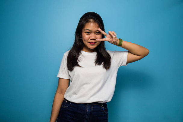 Portrait of Young beautiful asian women using white T-shirt with blue isolated background, showing peace sign and smiling, Victory sign, two sign - Valokuva, kuva