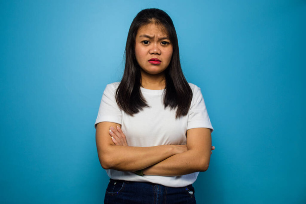 Portrét mladé krásné asijské ženy pomocí bílého trička s modrým izolovaným pozadím, skládací ruce a mrzutý - Fotografie, Obrázek