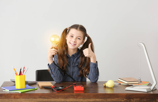Cute little schoolgirl with glowing light bulb at table against grey background - Photo, image